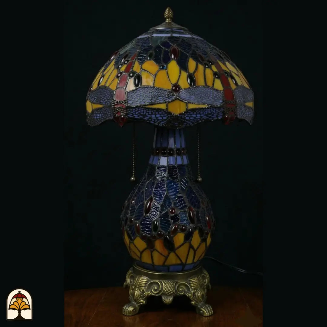 Onverlichte Tiffany tafellamp Libellula Azurea ⌀30 cm op donkere achtergrond – koepelkap met libellenmotief in blauw en amber glas-in-lood, glazen bolvoet met trekschakelaars