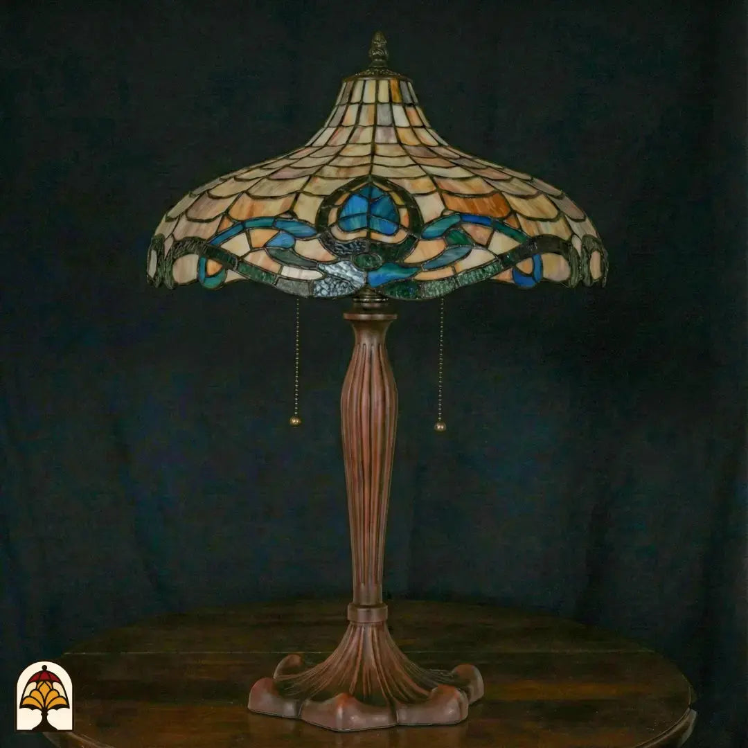 Onverlichte Tiffany tafellamp op donkerblauwe achtergrond — brede schaalvormige kap met amber schubpatroon, turquoise/blauwe rand met lotusmotief, bronzen voet met trekschakelaars