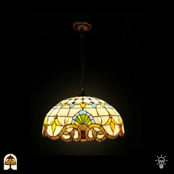 Verlichte Tiffany hanglamp Barok ⌀40 op donkere achtergrond, koepelvormige kap met crème glas-in-lood panelen, gele tulpmotieven, blauw-groene waaier, gouden barok rolwerkrand onderaan, rode juweel centraal, bronzen ketting en plafondrozet