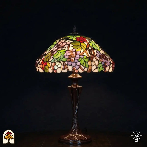 Verlichte Tiffany tafellamp Sylva Berria met brede koepelvormige kap, wijnrankmotieven met rode en lila druiventrosjes en groene bladeren in glas-in-lood, bronzen Art Nouveau vaasvoet met trekschakelaars, donkere achtergrond