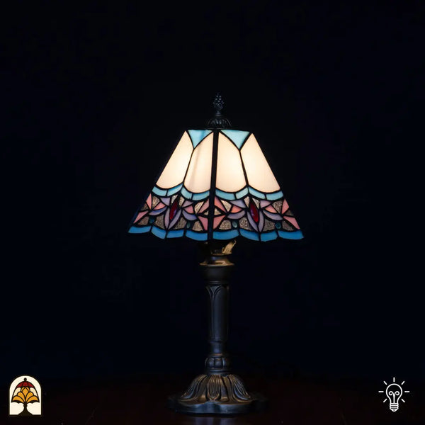 Verlichte Tiffany tafellamp met trapeziumvormige kap, witte/crème bovenpanelen, lichtblauwe randen, roze lotusbloemen en rode accenten onderaan, klassieke kandelaarvoet, zwarte achtergrond