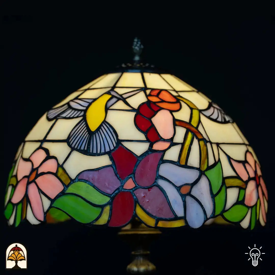 Close-up verlichte kap Tiffany tafellamp Colibrì — crème glas met kolibrie (gele/blauwe vleugels), rode en oranje bloemen, paarse en blauwe bloemen onderaan, groene bladeren