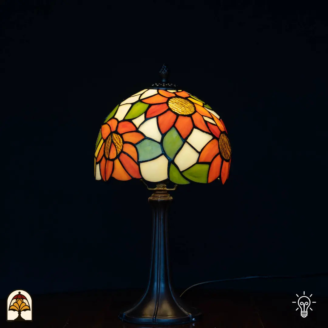 Tiffany tafellamp Solea verlicht op zwarte achtergrond — crème/witte koepelkap met oranje zonnebloemen en gele centra, groene bladeren, slanke bronzen Art Nouveau voet