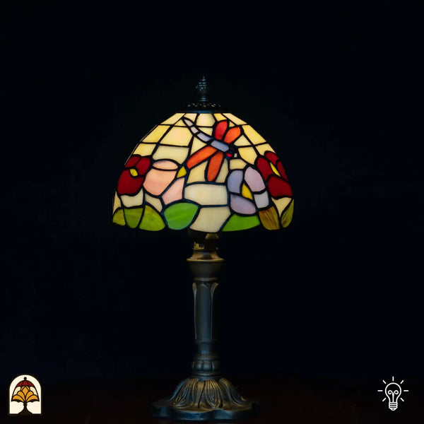 Verlichte Tiffany tafellamp met koepelvormige kap met oranje/rode libelle, rode bloemen, roze en paarse bloemen en groene bladeren op crème/gele achtergrond, klassieke kandelaarvoet, zwarte achtergrond
