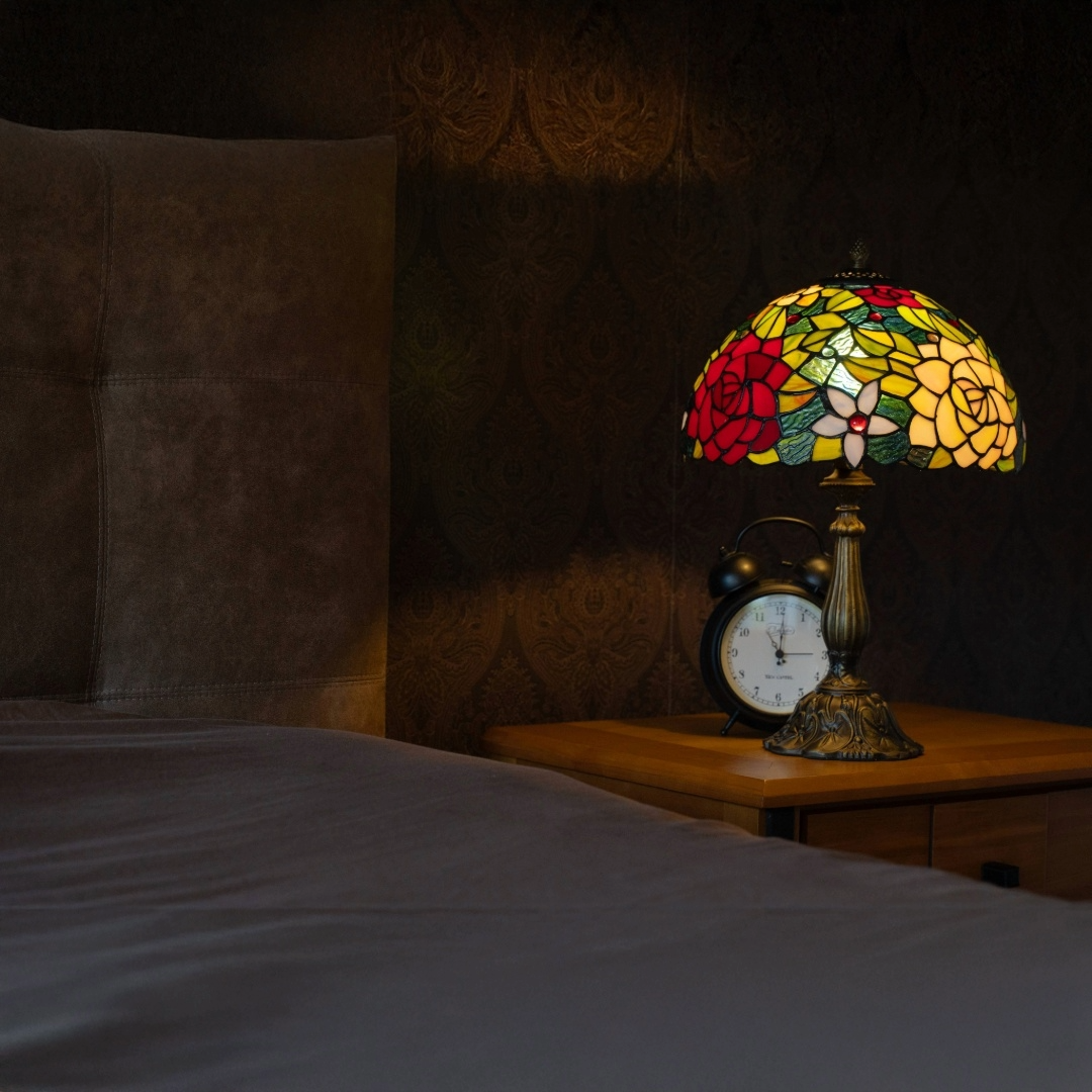 Een Tiffany tafellamp met een kleurrijke glas-in-lood kap vol bloemmotieven in warme tinten rood, geel en groen. De lamp staat op een sierlijk bronzen voet op een houten nachtkastje naast een klassieke wekker en verspreidt een zacht, warm licht dat de kamer een knusse, nostalgische sfeer geeft.