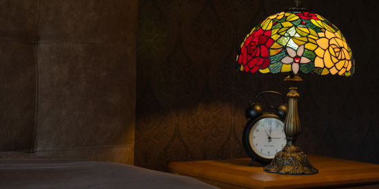 Een Tiffany-tafellamp met een kleurrijke glas-in-lood kap vol bloemmotieven in warme tinten rood, geel en groen. De lamp staat op een sierlijk bronzen voet op een houten nachtkastje naast een klassieke wekker en verspreidt een zacht, warm licht dat de kamer een knusse, nostalgische sfeer geeft.