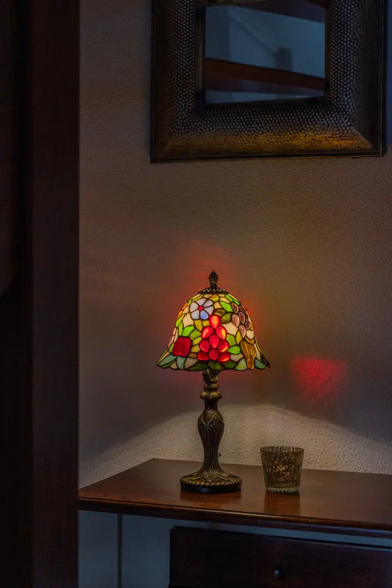 Kleurrijke Lamps by Tiffany tafellamp van glas-in-lood op een houten kastje, warm brandend en een zachte sfeergloed werpend tegen de muur.