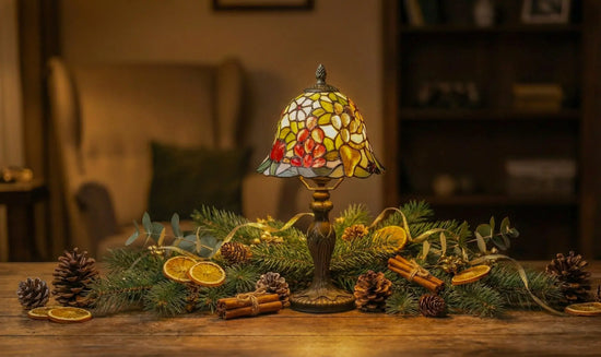 Tiffany tafellamp met gekleurde glas-in-lood kap op een houten tafel, omringd door dennentakken, dennenappels, gedroogde sinaasappelschijfjes en kaneel, in een warm verlichte woonkamer.