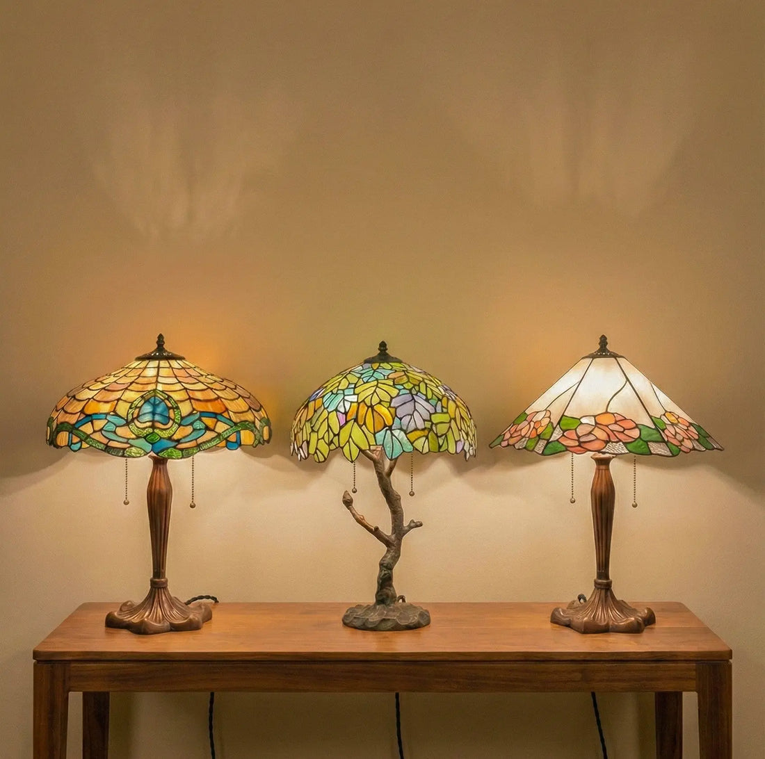Drie Tiffany tafellampen in verschillende stijlen op een houten tafel, met warm gekleurd glas-in-lood en sfeervol licht tegen een rustige wand.