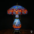 Tiffany lamp tafellamp verlicht, koepelvormige kap met blauw glas-in-lood bovenzone, oranje-rode middenzone met blauwe en amber glaskralen, lavendelblauwe libelle rand onderaan; peervormige lichtblauwe voet en bronzen leeuwenpootvoet
