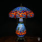 Tiffany lamp tafellamp verlicht, koepelvormige kap met blauw glas-in-lood bovenzone, oranje-rode middenzone met blauwe en amber glaskralen, lavendelblauwe libelle rand onderaan; peervormige lichtblauwe voet en bronzen leeuwenpootvoet