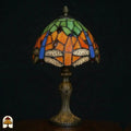 Onverlichte Tiffany tafellamp Libellula Azul ⌀20 op donkere achtergrond, oranje kap met groene bovenpanelen, blauwe libellenvleugels en libellenfries, rode en groene juwelen, bronzen Art Nouveau voet
