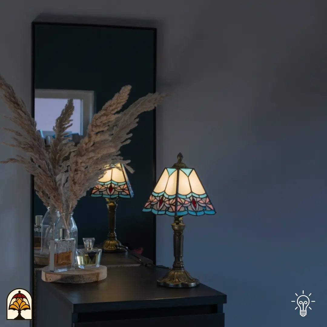 Lifestyle — verlichte Tiffany tafellamp op zwart dressoir voor grijze muur, spiegel met reflectie, pampasgrassen en parfumflesjes ernaast