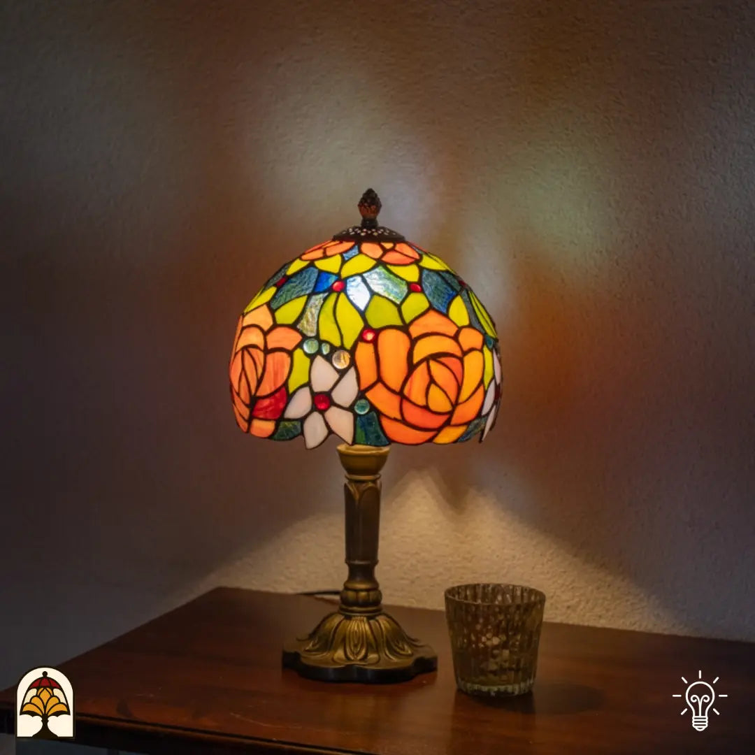 Tiffany tafellamp Fiore verlicht op plank naast witte muur — warm licht op achtergrond, glazen theelichtje rechts naast de lamp