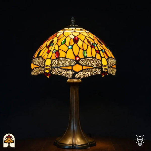 Verlichte Tiffany tafellamp Libellula Aurea ⌀30 op donkere achtergrond, koepelvormige kap met goudgele glas-in-lood panelen, libellenfries onderaan met beige vleugels, rode en groene ovale juwelen, bronzen trompetvoet