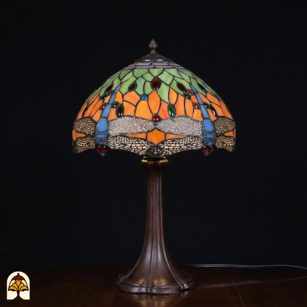 Onverlichte Tiffany tafellamp Libellula Azul ⌀30 op donkere achtergrond, koepelvormige kap met oranje panelen, groene bladmotieven, blauwe libellenmotieven en blauwe glasaccenten, rode en groene ovale juwelen, bronzen trompetvoet