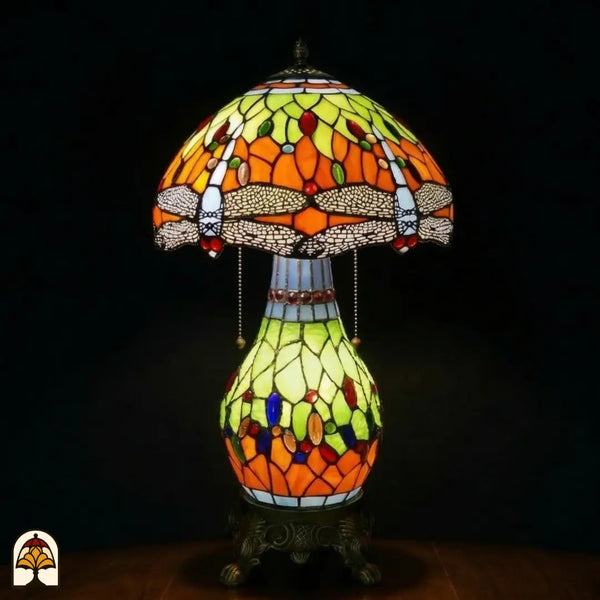Verlichte Tiffany tafellamp Libellula Lumina ⌀30 op donkere achtergrond, koepelvormige kap met oranje en geel-groene panelen, libellenfries, bolvormige glas-in-lood voet met geel-groene en oranje panelen en gekleurde juwelen, bronzen leeuwenpootvoet, trekschakelaars