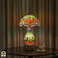 Verlichte Tiffany tafellamp Libellula Lumina ⌀30 op ronde houten tafel voor lichtbeige gordijnen, glas-in-lood kap en -voet volledig verlicht, theekopje op schotel naast de lamp