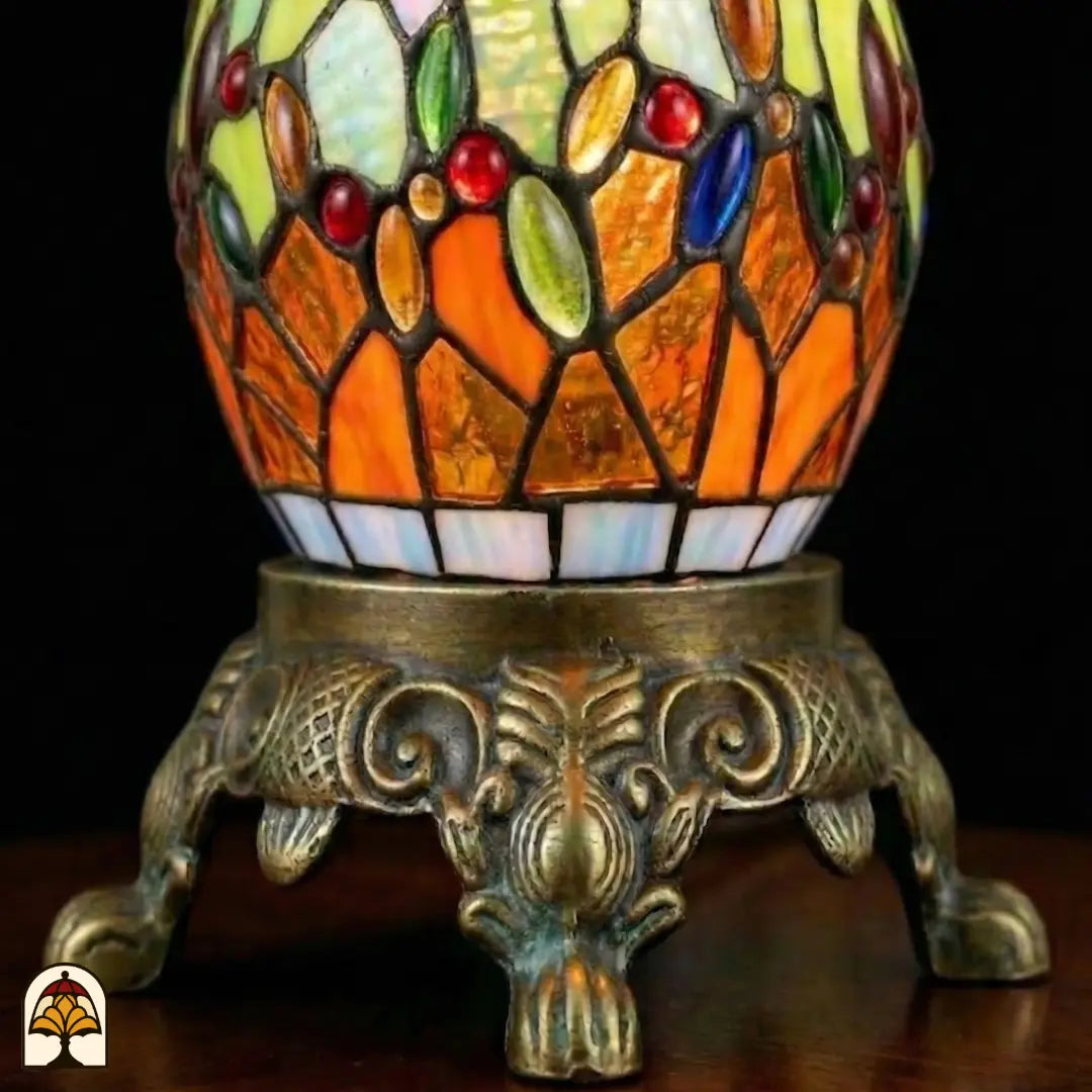 Close-up bronzen siervoet Libellula Lumina ⌀30 met glas-in-lood bolvoet, oranje en groene panelen met gekleurde juwelen, bronzen voetstuk met acanthusblad- en krulmotieven en leeuwenpoten