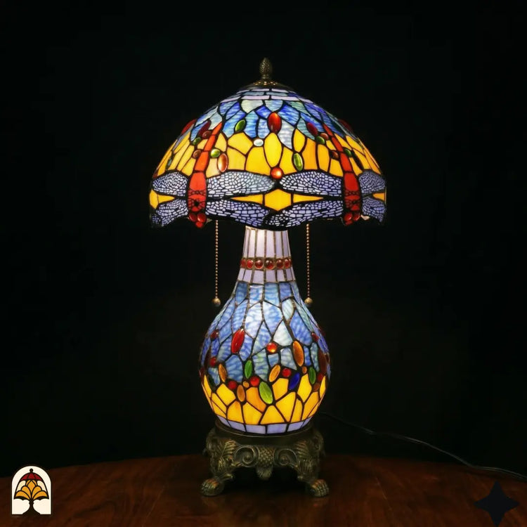 Verlichte Tiffany tafellamp Libellula Azurea ⌀30 op donkere achtergrond, koepelvormige kap met gele en blauwe panelen, rode libellenfries, gekleurde juwelen, bolvormige glas-in-lood voet met lichtblauwe en gele panelen, bronzen leeuwenpootvoet, trekschakelaars