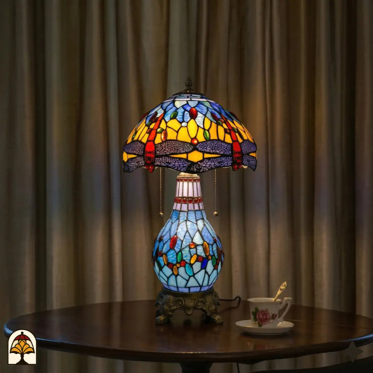 Verlichte Tiffany tafellamp Libellula Azurea ⌀30 op ronde houten tafel voor lichtbeige gordijnen, kap en glas-in-lood voet volledig verlicht, theekopje op schotel naast de lamp