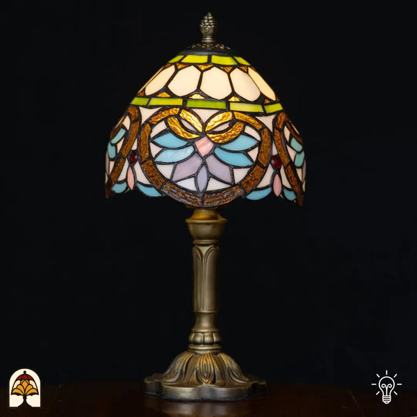 Verlichte Tiffany tafellamp Lucette ⌀20 cm op zwarte achtergrond – crème koepelkap met groene sierband, gouden libelle-/bladvormen, lichtblauwe en roze bloemblaadjes, rode edelstenen, bronzen kolomvoet