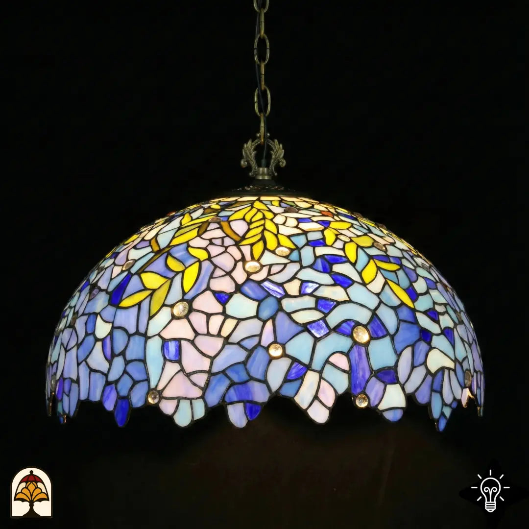 Onverlichte Tiffany hanglamp Drizzle Bleu ⌀50 op donkere achtergrond, koepelkap met lila-blauwe en turquoise glas-in-lood panelen, gele bladmotieven, ronde kristalaccenten, geschulpte onderrand, bronzen ketting