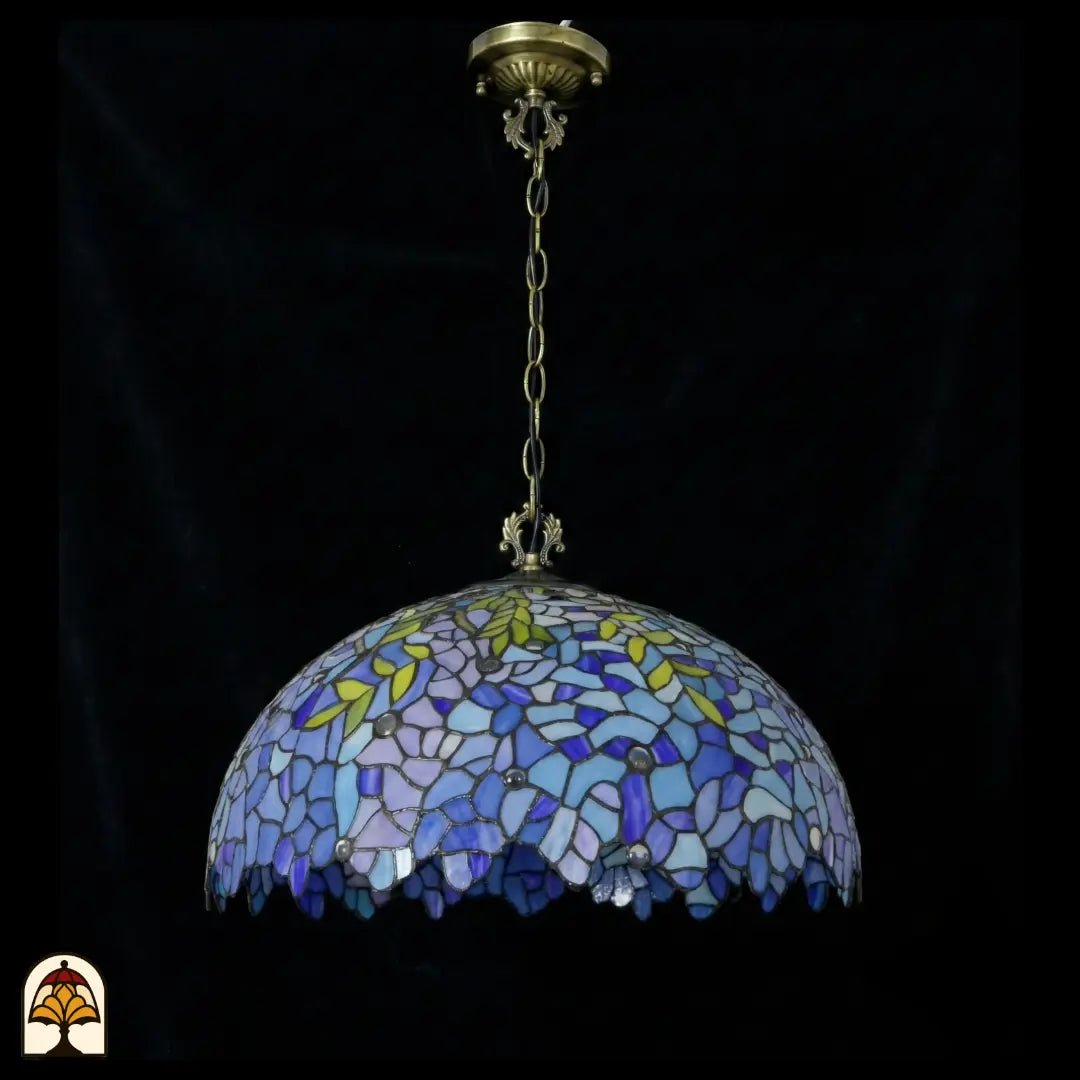 Onverlichte Tiffany hanglamp Drizzle Bleu ⌀50 op donkere achtergrond, blauwe en paarse koepelkap met gele bladaccenten en geschulpte onderrand, bronzen ketting met plafondrozet