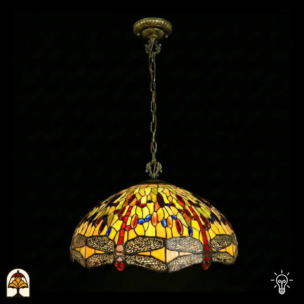 Onverlichte Tiffany hanglamp Libellula Aurelia ⌀50 op donkere achtergrond, grote koepelvormige kap met gele glas-in-lood panelen, libellenfries onderaan, rode en blauwe juwelen, bronzen ketting en plafondrozet