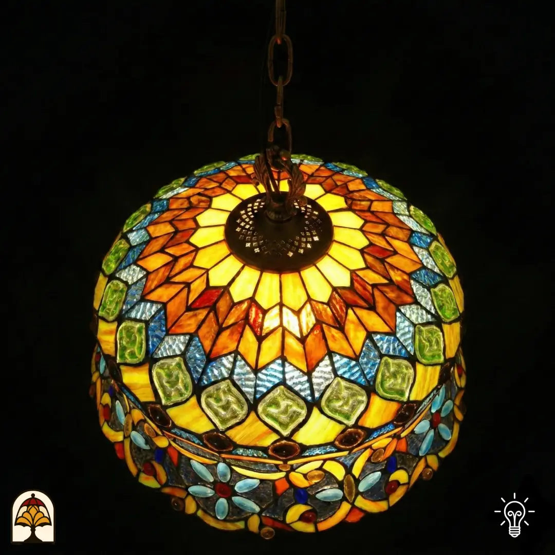 Bovenaanzicht van tiffany lamp hanglamp verlicht, stralend patroon met amberkleurige vlakken, groene glaskralen en blauwe mozaïekvakken, bronzen ventilatieplaat zichtbaar in het midden