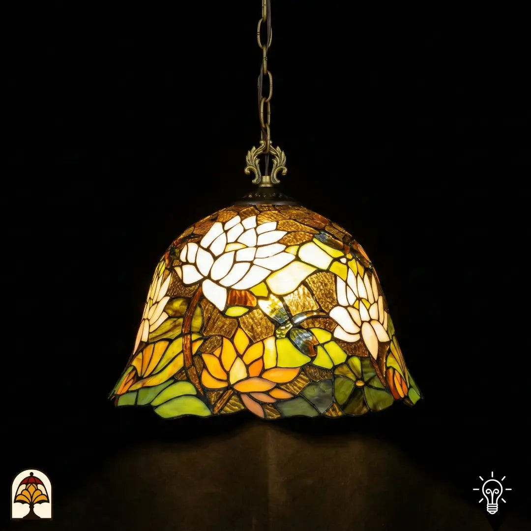 Tiffany lamp hanglamp verlicht van dichterbij, klokvormige kap met witte lotusbloemen bovenaan, oranje-gele lotusbloemen onderaan, groene waterleliebladen en blauwe libelle op bruinamberen achtergrond, opgehangen aan bronzen ketting