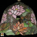 Detailopname van tiffany lamp klokvormige kap onverlicht met roze lotusbloem centraal, oranje lotusbloemen onderaan, groene waterleliebladen en een bruinoranje libelle met blauwe details