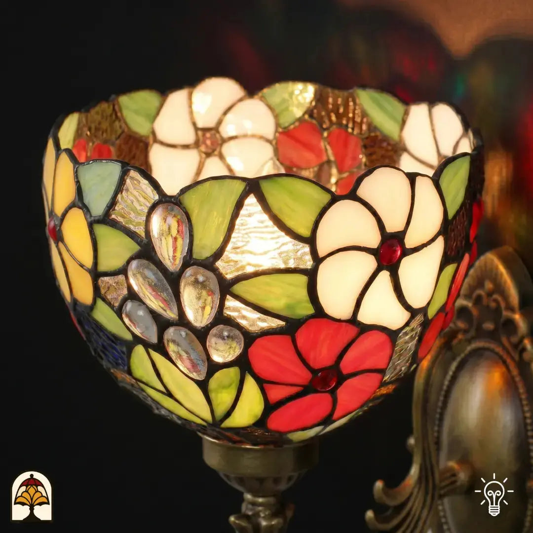 Detailopname van tiffany lamp wandlamp glas-in-lood kap verlicht, komvormige kap met witte bloemen, rode bloemen, groene bladeren en druppelvormige transparante glaskralen op bruinamberen achtergrond, bronzen fitting zichtbaar