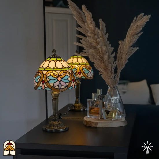 Tiffany tafellamp met bloemmotief in zachte pastelkleuren, geplaatst op een donkere kast naast een glazen vaas met pampasgras en parfumflesjes, straalt warm licht uit in een sfeervolle slaapkamer.

