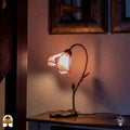 Een sfeervolle Tiffany tafellamp in de vorm van een zachtroze bloemkelk, gemonteerd op een sierlijke bronskleurige voet met een knopje en bladmotief. De lamp staat op een houten sidetable en verspreidt een warm, roze licht tegen een witte wand. Op de achtergrond hangen twee vierkante, gedecoreerde spiegels naast een houten trappartij.