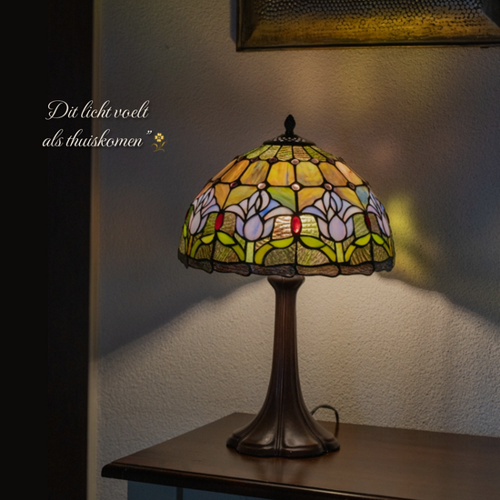 Tiffany tafellamp met handgemaakte kap in bloemmotief met groene bladeren en rode accenten. Plaatsing op een houten kast, ideaal als sfeerlamp die warmte en karakter in huis brengt.