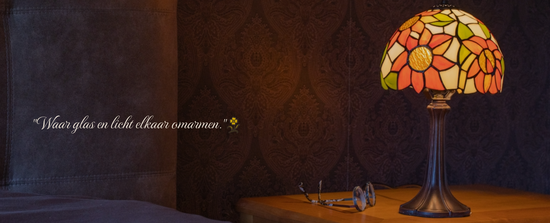 Authentieke Tiffany tafellamp met handgemaakte kap in zonnebloemmotief, in warme oranje en gele kleuren. Plaatsing op een houten nachtkastje met bril, ideaal als sfeervolle slaapkamerlamp.