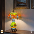 Verlichte Tiffany tafellamp Libellula Lumina ⌀30 cm in interieur – oranje en geel-groene glas-in-lood kap met libellenmotief, bronzen leeuwenpootvoet, warm licht