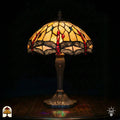 Verlichte Tiffany tafellamp Libelle Elegance met koepelvormige geel-amber kap, rode druppelvormige en blauwe ronde glas-in-lood accenten, libellenmotieven langs de rand, bronzen geribbelde vaasvoet, donkere achtergrond