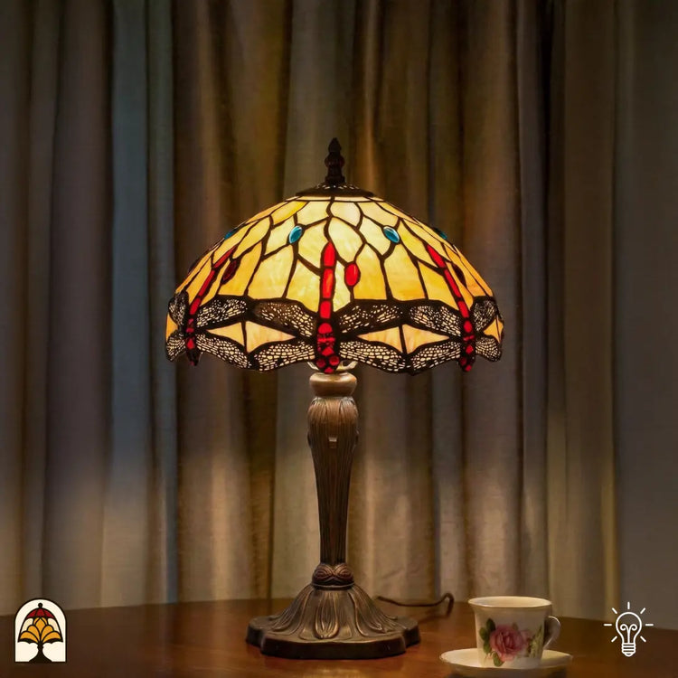 Verlichte Tiffany tafellamp Libelle Elegance op houten tafel voor gordijnen, geel-amber kap met rode accenten en libellenmotieven, theekopje ernaast