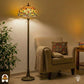 Tiffany vloerlamp Libellula Aurea verlicht in woonkamer — naast beige fauteuil met deken en kussen, groene plant links, bijzettafel met boeken rechts