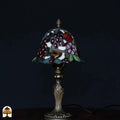 Onverlichte Tiffany tafellamp op donkere achtergrond — klokvormige kap met bordeaux druiven, amber glas, roze en blauwe bloemen, groene bladeren, klassieke kandelaarvoet