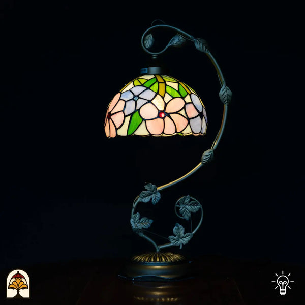 Verlichte Tiffany tafellamp met gebogen zwaan-hals arm, koepelvormige kap met roze bloemen en groene bladeren op crème achtergrond, donkere bladervormige voet met ronde basis, zwarte achtergrond
