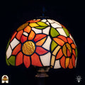 Close-up verlichte kap Tiffany tafellamp Solea — oranje zonnebloembladen met goud/bruine centra, groene bladeren, crème/witte vlakken