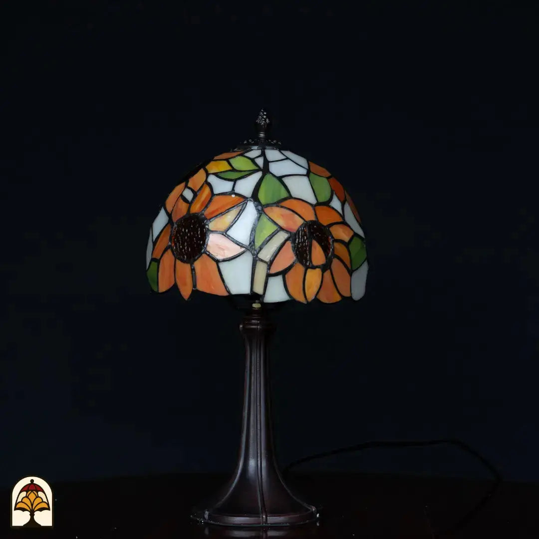 Tiffany tafellamp Solea onverlicht op donkere achtergrond — kap met oranje zonnebloemen en blauwe/witte vlakken, groene bladeren, slanke Art Nouveau voet