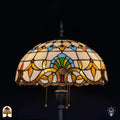 Close-up verlichte kap Tiffany vloerlamp Barok — crème/amber glas, centrale blauwe boog met groene waaier, rode edelsteen op bronzen rand, trekschakelaar