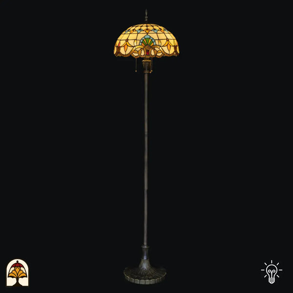 Tiffany vloerlamp Barok verlicht op zwarte achtergrond — barokse koepelkap crème/amber met blauwe, groene en gele motieven, bronzen rand met trekschakelaar, lange paal met brede voet