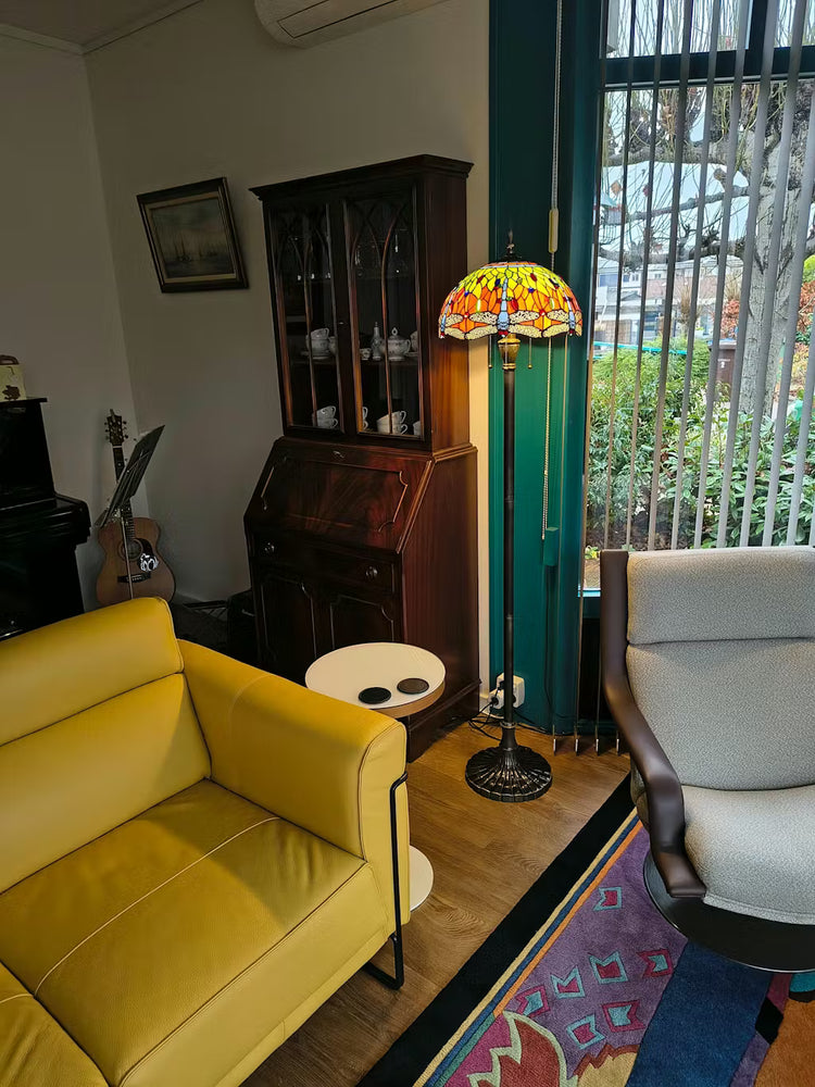 Tiffany vloerlamp met geel-oranje glas-in-lood kap en libellemotief, zorgt voor warm sfeerlicht in de woonkamer.